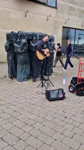 A young man with dark hair is playing an acoustic guitar and singing while standing in front of a large, dark gray sculpture of cloaked figures. He is wearing a black hoodie with white initials "JS" and dark pants. An open guitar case, a black amp, a water bottle, and a small red cart are positioned near him on the paved ground. People are walking in the background. The building behind the sculpture appears to be made of tan stone. The camera moves slightly, providing different angles of the scene.