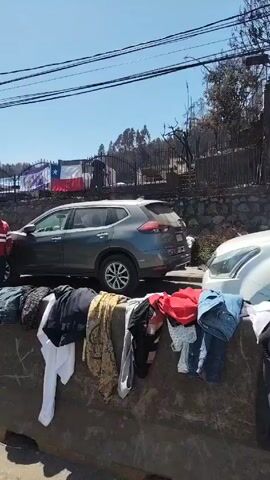 The video shows a street scene with various activities taking place in front of a fenced-off area. In the initial shot, two men in red and white striped shirts are standing by the roadside with large containers of liquid. A gray SUV is parked near them. A few vehicles including a white SUV pulling a red trailer pass by. A black dog also crosses the street. Further along the road, a large red dump truck drives by, and there are remnants of what appear to be garments or textiles on the barrier. The backdrop includes a clear blue sky, utility poles, and a hillside with visible signs of fire damage, indicating a post-fire environment. A Chilean flag is visible on the other side of the fence.
