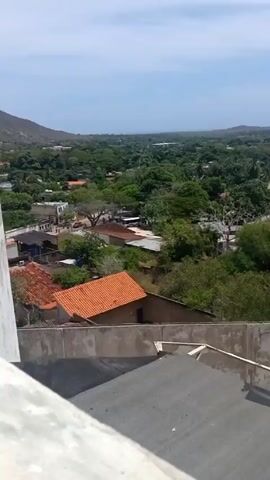 From an elevated, possibly rooftop position, the video shows an expanse of green vegetation interspersed with residential buildings featuring predominantly red-tiled roofs. The architecture is low-rise. In the distance, a coastline is partially visible beneath a partly cloudy sky.  The camera pans slightly to reveal a large, semi-circular structure, possibly an auditorium or covered arena, before returning to its original position. The scene appears to depict a small town or rural community.