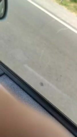 The video opens with a close-up view of a car's interior door panel, showing a window lock button and part of a door handle. The perspective then shifts to an exterior view of a paved road with a white line, bordered by a field with dry grass and bushes. Further along the road, a black SUV drives past, its windows tinted. Sunlight indicates it is daytime.