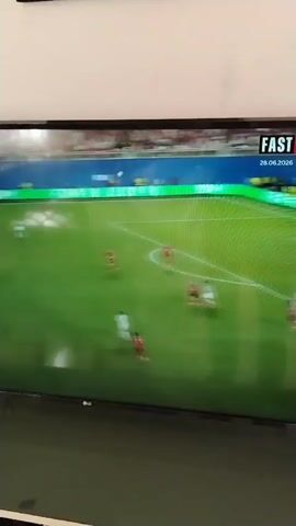 A television screen displays a soccer match. The field is green, with players in white and red uniforms scattered across it. A scoreboard in the upper right corner shows "FAST" and the date "28.06.2020". Spectators can be seen in the background, filling the stadium seating. The image is slightly blurred, suggesting it was taken from an angle or with a moving camera.