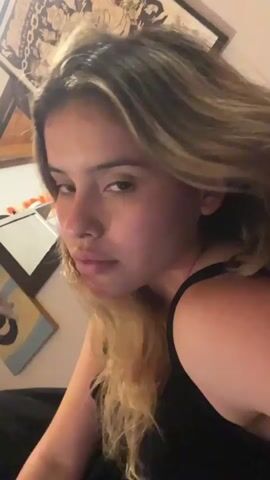A close-up video shows a young woman with blonde-streaked brown hair wearing a black top. She is lying down and looking into the camera with a neutral expression. The background is blurred but appears to be a room with framed artwork on the wall. The lighting is dim.