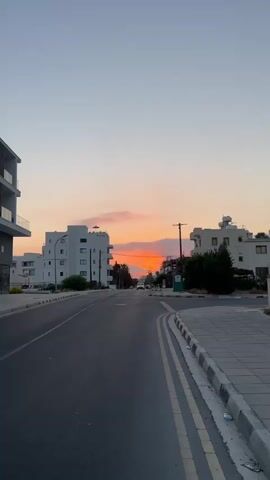 A vertical video captures a street view during sunset. The sky displays hues of orange and pink as the sun dips below the horizon. Modern white buildings with balconies line both sides of the asphalt road. The road has double yellow lines and a paved sidewalk with stone tiles. A few cars are visible on the road, and streetlights are present. The overall scene depicts a quiet, urban environment at dusk.