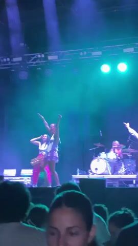 This video shows a live music performance on a stage. Three women are visible on stage. One woman in the center is singing and dancing. Another woman to her left is playing a guitar. The third woman to her right is playing a saxophone. The stage is lit with green and blue lights, and there is smoke or fog effects. In the foreground, the backs of several audience members are visible.