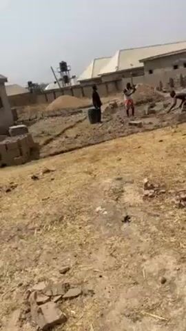 Three individuals are working at a construction site. One is near a black barrel, possibly supervising. Another is swinging a pickaxe to break apart construction blocks. The third is bent over, possibly placing blocks or clearing debris. The site appears to be an open field with some dirt, grass and scattered brick remnants. In the background, there are buildings, including one that appears partially constructed, and a water tower. The weather seems to be sunny and clear.