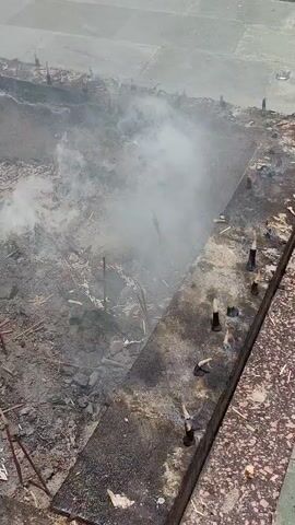 The video captures an aerial view of an outdoor scene characterized by debris, smoke, and what appear to be the remnants of some sort of structure or pyre. The ground is covered in ash, charred wood, and scattered objects. A significant amount of white smoke is billowing from the center of the scene, obscuring some of the details. Several thin, stick-like objects, possibly incense sticks or remnants of construction material, are visible protruding from the ground at various angles, particularly in the foreground and mid-ground. In the foreground, a dark, flat surface with several pointed objects embedded in it runs along the right side of the frame, potentially a protective barrier or part of a larger structure. The overall impression is one of destruction or a recent burning event. There are no people visible in the footage. The lighting suggests it is daytime.