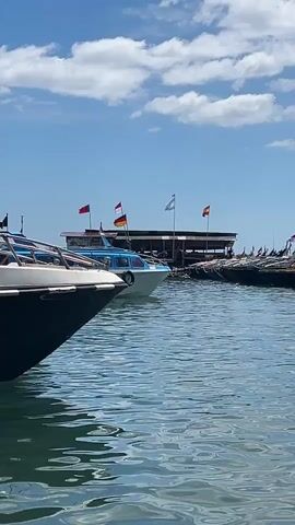 The video shows a harbor with several boats docked. The water is calm with small ripples. In the foreground, the bow of a dark-colored boat is visible on the left. Further back, a light blue and white boat is docked between other boats. Above the docked boats, a building with a roof is visible. Several flags are flying from poles on or near the building, including what appears to be the flag of Germany, and possibly others. More boats are visible in the background, stretching further down the harbor. The sky is blue with scattered white clouds. The scene is well-lit, suggesting it is daytime. There are no people clearly visible in the frame.