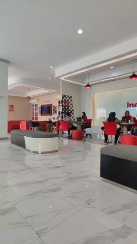 Here is a matter-of-fact description of the video:

The video shows the interior of an IndiHome by Telkomsel office or customer service center. Several small round tables, surrounded by red chairs and stools, are scattered throughout the room. People are seated at these tables, some with laptops, appearing to be engaged in conversations. The space is modern, with white marble tile flooring, gray and black benches, and pendant lights with red shades. A large IndiHome by Telkomsel logo is prominently displayed against a white wall in the background.