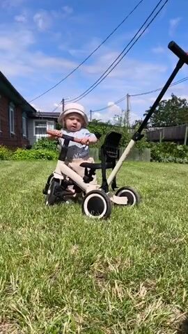 A child wearing a hat and a light-colored shirt and pants sits on a beige tricycle with a push handle. The child is in a grassy backyard with a wooden house in the background. The sky is blue with some clouds. The child's hands are on the handlebars. The tricycle has three wheels with white rims. The child is looking to the left.