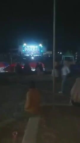 The video shows a nighttime outdoor event with a stage illuminated by bright lights in the background. A crowd of people is gathered in front of the stage, many sitting on the ground or on low structures. In the foreground, several individuals are also seated, facing away from the camera and towards the stage. Umbrellas, tents, and possibly food stalls are visible scattered throughout the venue. A tall pole stands prominently in the middle ground, slightly obscuring the view of some of the audience members. The overall atmosphere suggests a concert, festival, or public gathering.
