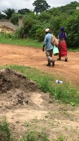Two individuals are walking away from the camera down a dirt road. The person on the left is wearing a light blue long-sleeved shirt, khaki pants, a pink cap, and dark boots. They are carrying a light brown sack over their left shoulder. The person on the right is wearing a pink long-sleeved shirt, a purple headscarf, and red pants. They are carrying a light green bucket with their right hand. Both individuals are walking on a reddish-brown dirt path. The path is bordered by green grass and vegetation on both sides. In the background, there is a pile of sand or building material, a low wall, and trees. A plastic water bottle is lying on the grass to the right of the path. The sky appears overcast. The ground in the foreground on the left is a pile of dark soil.