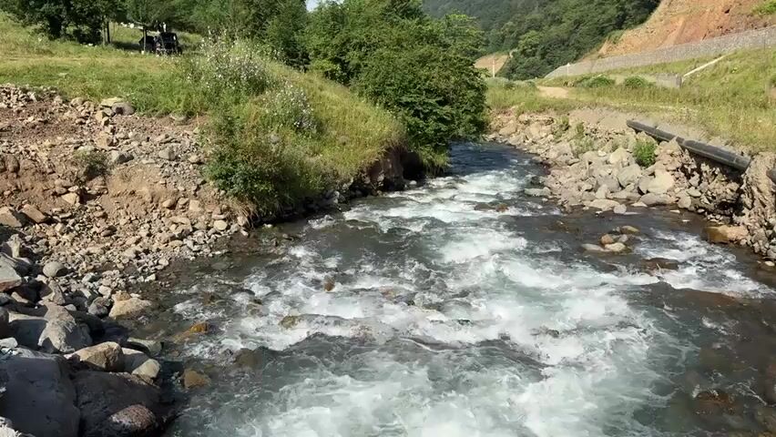 The video shows a fast-flowing river with white water cascading over rocks. The riverbanks are lined with stones and some patches of grass and small bushes. In the background, there are trees and a sloping hillside with exposed soil and some construction, possibly a road or terracing, with a pipe running along the embankment on the right side of the river. The sky is clear and blue, suggesting a sunny day. There are no people visible in the frame.