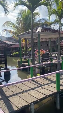 A video pans across an outdoor dining area with thatched-roof gazebos over a body of water. Palm trees and other foliage are visible, as well as a telecommunications tower in the distance. A wooden walkway with purple and green railings leads through the area. The sun is shining, and there are clouds in the sky. There are no people visible in the video.