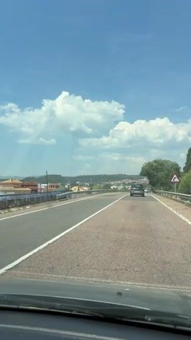 The video depicts a car journey on a two-lane highway. The road is asphalt, marked with a solid white line, and has metal guardrails. A car drives in the same direction, in the distance. The background features a town nestled among hills, under a bright blue sky with puffy white clouds. The camera is inside the moving vehicle. A traffic sign can be seen on the right side of the road, as well as trees.