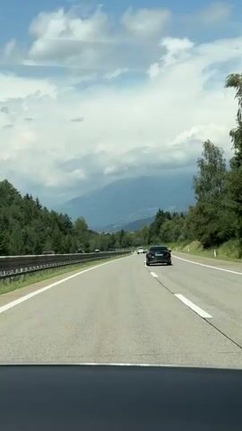 A first-person view from a vehicle traveling on a multi-lane highway. The highway is paved with asphalt and marked with white lines. Trees and greenery line the sides of the road. In the distance, mountains are visible under a cloudy sky. Several other vehicles are on the highway, including a black car in front of the viewer's vehicle. The black car has its brake lights on, indicating it is slowing down. The video appears to be filmed on a bright, possibly sunny day.