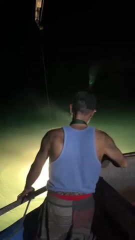 A man wearing a blue tank top and patterned shorts stands on a boat at night, looking away from the camera. He is holding onto the side of the boat. In the background, a net is being pulled out of the dark water by a crane or similar mechanism. A bright light is illuminating the water below the net.