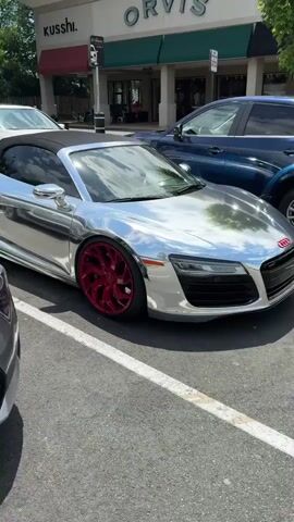A silver Audi R8 Spyder is parked in a parking space in front of a shopping center. The car has red wheels and red Audi logo on the hood. Other cars are parked in the background, and stores and restaurants are visible in the distance. The sky is partly cloudy.
