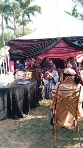 The video shows a gathering of people outdoors, seemingly at an event under a large tent adorned with black and pink fabric. Many are seated on chairs, some with traditional attire. A woman in a white outfit is holding a baby, and a small child is standing nearby. A table covered with a black cloth is in the foreground, displaying some items and decorative elements. Palm trees are visible in the background, suggesting a tropical or subtropical environment. The atmosphere appears social and perhaps celebratory, though details about the specific occasion are not explicitly shown.