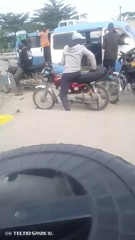 The video shows an outdoor scene with several motorcycles and a white van. A person in a gray hoodie and dark pants is sitting on a red motorcycle, facing away from the camera. Another person wearing a black jacket and dark pants is standing near the van. There are other people and motorcycles visible in the background, as well as a road with some traffic. The ground appears to be dirt or sand. The overall impression is of a busy outdoor area, possibly a transportation hub or marketplace.