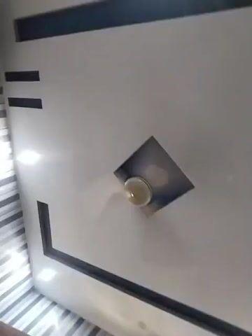 The video displays a ceiling, decorated with a minimalist design. The base color of the ceiling is white, and it's contrasted with geometric shapes in dark shades, including a long rectangle, short parallel lines, and a square, tilted to form a diamond. Inside the diamond shape, there's a light fixture with a translucent globe. To the right of the ceiling, a portion of a curtain with a floral pattern is visible.