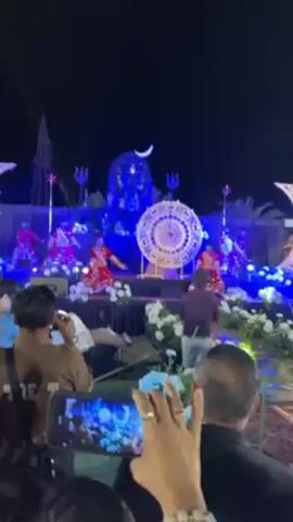 Several young dancers in matching red and gold outfits perform on a stage at an outdoor event at night. Behind them is a large statue of a deity's head, decorated with a crescent moon and tridents. A large, ornate circle hangs in the background. The stage is lit with blue and white lights, and there are decorations of white flowers. Several attendees are visible, including people holding up cell phones to record the performance. The camera shakes slightly while filming the dance.