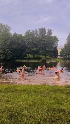 A group of people are swimming and playing in a lake. The water is murky brown and shallow enough for people to stand. The area is surrounded by lush green grass and trees. In the background, there appears to be a building and a cloudy sky. Some individuals are splashing, while others are wading or sitting in the water. One person is visible in the process of diving or jumping into the water from the right side of the frame.