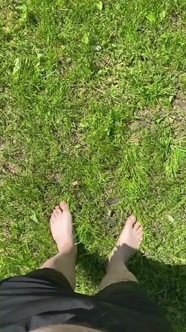A first-person point of view video shows a person walking barefoot outdoors on a sunny day in Suzdal, Russia. The person is wearing black shorts. They step from a patch of vibrant green grass onto a grey brick pathway, which has some weeds growing between the bricks. The sun casts shadows, indicating bright daylight.