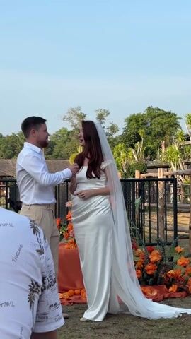 The video captures a joyous moment at an outdoor wedding ceremony. A man, dressed in a white button-down shirt and beige trousers, stands facing a woman in a white, satin-like wedding gown with a high slit and a long, flowing veil. The setting appears to be a grassy area, with vibrant orange and yellow floral arrangements behind the couple. In the background, there's a black metal fence and lush green trees under a clear blue sky, suggesting a warm, sunny day.

At the beginning of the video, the couple is holding hands, with the man gently touching the woman's chin or cheek. As applause and cheers erupt from off-screen, they raise their joined hands above their heads in a celebratory gesture. Immediately after, they embrace and share a passionate kiss, with the woman lifting one leg slightly off the ground. The kiss is held for a few seconds as the crowd's enthusiastic cheers and applause grow louder. A person in the foreground, partially visible, is wearing a white shirt with text, parts of which read "THAILAND" and "AND BEACH." The overall atmosphere is celebratory and full of happiness.