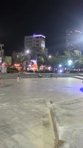At night, a wide, paved urban plaza is lit by overhead streetlights. Several children are playing, some running and kicking a ball.  Across the street, multiple businesses and hotels are visible, including "The Beach Hotel" and "Luna Hotel" with their signs illuminated. Cars are parked on the street, and palm trees line the plaza. The overall scene is a public space where children are freely playing under the artificial light of the city.