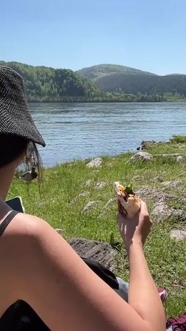 The video captures a peaceful outdoor scene by a body of water in Sliznevo, Russia, on a sunny day.

The first shot shows a person, viewed from behind, wearing a black hat and sitting on a grassy bank beside a wide, calm river or lake. The person holds a wrap or shawarma in their hand, seemingly enjoying a meal outdoors. In the background, forested hills rise across the water under a clear blue sky.

The camera then zooms in on the person's back, revealing a tattoo on their left shoulder. The tattoo features an abstract design incorporating a crescent moon, a vertical line, and geometric symbols.

The final shot focuses on a duck, likely a mallard, walking and pecking at the grass right at the water's edge. The clear blue water and green grass indicate a pleasant, warm day.
