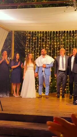 The video shows a wedding ceremony under a chuppah adorned with string lights. A bride in a white gown and a groom in a blue suit stand center, holding hands. To the bride's left, two women in formal dresses clap, and to the groom's right, two men in suits stand, one with his mouth open. The groom wears a tallit over his suit. The scene is set on a wooden platform with trees and foliage visible in the background.