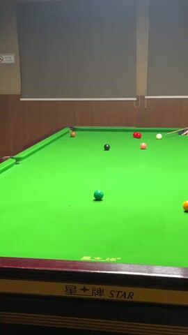 A snooker player is aiming at a green ball on a snooker table. The table is covered with green felt and has red, pink, white, and orange balls on it. The player is wearing a grey shirt. The walls are wood-paneled and there is a blind on the wall.