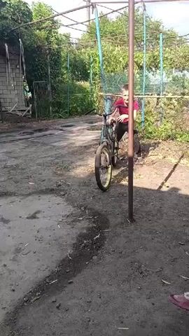 A boy on a bicycle is seen in an outdoor area. He is wearing a red shirt and dark pants. The boy is riding the bicycle towards the camera. He is holding onto the handlebars and appears to be pedaling. The bicycle has a black frame and yellow rims. In the background, there is a building and some vegetation. The ground is dirt and gravel. There is a pole in the foreground, to the right of the bicycle. The sun is shining, casting shadows.