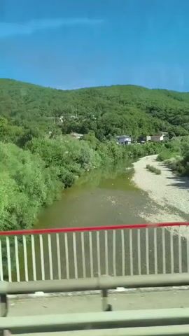 The video captures a scenic view from what appears to be a bridge or elevated walkway. In the foreground, the railing of the walkway is visible with white vertical bars and a red top rail. Three people are walking across the walkway, one on the left wearing a white hat and a light-colored top, another in the center also with a white hat and a light top, and a third person on the right wearing a white hat and a light-colored top. Beyond the walkway, a river flows, with green trees lining its banks and a rocky shore on the right side. In the background, rolling green hills covered with lush trees rise up. A few houses are scattered among the trees on the hillsides, and the sky above is a clear blue. The video pans slightly to the right, revealing more of the landscape.