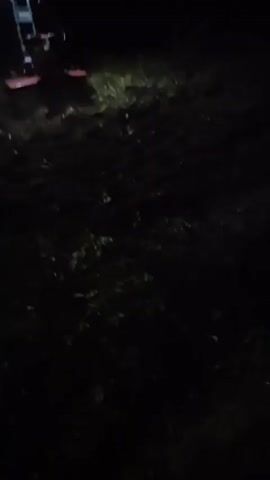 A person wearing a headlamp is operating a rototiller in a dark outdoor environment. The rototiller is red and blue and appears to be tilling soil. The ground is uneven and covered with some vegetation. The video is shaky and the lighting is poor, indicating it was likely filmed at night with limited light sources. The person is moving from left to right across the frame.