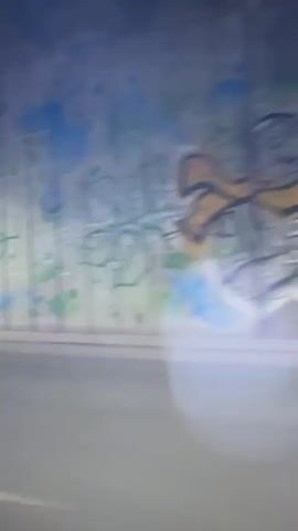 The video captures a blurred, fast-moving perspective from inside a vehicle, likely at night. The view is primarily of a road and a concrete wall or tunnel barrier passing by quickly. Light sources create lens flares, suggesting reflections on a window. The ground appears to be asphalt with painted lines. The environment transitions from a dark, possibly outdoor scene with distant lights to a more enclosed space, possibly a tunnel, with artificial lighting on the walls.