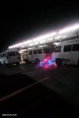 At night, the video shows a parking area with three white vans parked under a lighted shelter. The camera moves, initially focusing on the tail lights of a large vehicle with multiple red lights. The camera then pans to another vehicle with both yellow and green lights, briefly showing the dark outline of the vehicle itself.