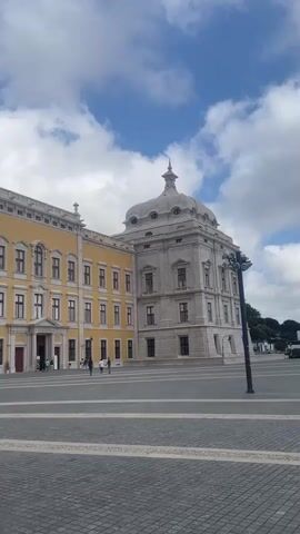 The video pans across a large, open square in front of an ornate, yellow building with a prominent white bell tower. Several people are visible in the distance, walking around the square and near the building entrances. The square is paved with a pattern of gray and white cobblestones. The sky is partly cloudy with patches of blue visible. The camera movement is smooth, suggesting a deliberate recording of the architecture and the surrounding area.