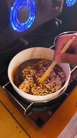 A close-up shot shows a bowl of instant ramen noodles being stirred with chopsticks. The noodles are a light brown color and are submerged in a dark, rich broth. The bowl is white and sits on a black stove. In the background, a device with glowing blue lights is visible. The noodles are being mixed vigorously by a hand holding wooden chopsticks. The lighting is dim, with a strong spotlight on the bowl of noodles, creating a dramatic effect. The overall impression is of someone preparing a hot meal.