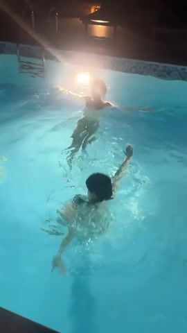 Two individuals are swimming in a brightly lit pool at night. The person closer to the camera, wearing a dark bikini top, initially has their head submerged before surfacing and treading water, creating splashes. The second person, further in the background, wearing a light-colored swimsuit, is seen floating on their back and then begins to swim away from the camera. A strong light source reflects off the water in the upper middle of the frame. A pool ladder is visible in the background.