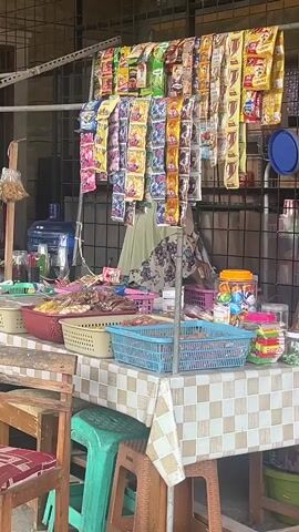 A street vendor stands behind a makeshift stall displaying an assortment of small packaged goods, likely snacks or instant food items, hanging from a metal rack. Below the hanging items, various baskets and containers hold more products. One person, wearing a hijab and a patterned top, is partially visible behind the stall, appearing to be tending to the merchandise. The stall is covered with a checkered tablecloth, and wooden and plastic stools are placed in front of it. The environment appears to be outdoors, with a tiled wall in the background.