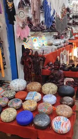 The video shows the interior of a souvenir shop filled with a wide array of items.  The foreground features a display table covered with red cloth, showcasing tongue drums in various colors and designs. Behind the drums are small statues of religious figures like Buddha and Hindu deities.  

The shop has checkered tile flooring and is filled with display tables covered with red cloth. These tables hold a variety of items including sculptures, dream catchers, and other decorative objects.  Shelves lining the walls display small figurines and other collectibles. The lighting is bright and the shop appears well-stocked. The camera slowly pans across the scene, offering a full view of the shop's interior.