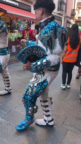 The video shows a group of people in colorful costumes dancing in a street. The costumes appear to be elaborate and embellished with sequins and metallic elements. Some of the costumes feature images of animals, such as lions. The dancers are moving in unison, with synchronized steps and arm movements. The background shows buildings and a few onlookers, suggesting a public event or celebration. The lighting indicates it is daytime.