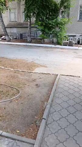 The video shows an outdoor scene. In the foreground, a paved walkway with a decorative pattern is visible, bordered by a concrete curb. To the left of the walkway, there is a patch of dirt and sparse vegetation. Beyond this, a paved area, possibly a road or parking lot, is seen. Further back, a building with multiple windows stands, partially obscured by trees. Construction materials, including pallets and stacks of what appear to be concrete blocks, are present near the building. A blue tractor with a yellow loader attachment is parked in front of the building. The lighting suggests it is daytime. The camera is positioned at a low angle, looking slightly upwards.