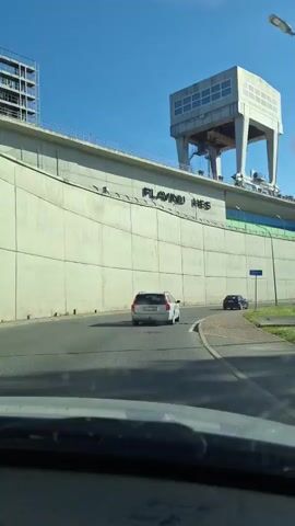 This is a vertical video clip filmed from a car, showing a drive along a road with a large concrete dam on the left. The dam is a significant structure with a distinctive tower-like building on top, featuring multiple glass windows. The word "SLAVINU" is visible, written in large, raised letters on the side of the dam. 

Two cars are driving on the road ahead of the camera, a silver station wagon in the left lane and a dark car in the right lane. The road has a paved surface with a distinct curb and a paved walkway with grass beyond it. The sky is a clear blue, indicating fair weather, and the sun is shining, suggesting it is daytime, likely late morning or afternoon.

The scene appears to be in an industrial or infrastructure setting, possibly related to hydroelectric power, given the dam's appearance. The overall mood is neutral, focusing on the view and the drive. There are no people prominently visible besides those in the cars. The clip provides a glimpse of the Aizkraukle hydroelectric power plant in Latvia.