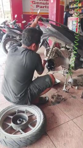A person is squatting on a tiled floor in what appears to be a motorcycle repair shop. The person, wearing a dark t-shirt and dark shorts, is facing away from the camera and is working on the rear section of a motorcycle. The motorcycle is partially disassembled, with parts on the floor around it. A detached motorcycle wheel with a tire is in the foreground. In the background, other motorcycles are parked, and shelves stocked with various items are visible on the wall. The overall impression is a busy workshop environment.