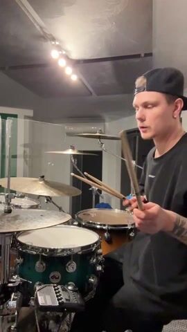 A young man is seen playing a drum set. The man is wearing a black shirt, a black cap, and has tattoos on his left arm. He throws the drumsticks up in the air and catches them. The drum set is green and silver. The environment is a recording studio, with a set of lights on the ceiling. There is also a glass partition visible in the background.