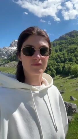 A woman with brunette hair pulled into a low ponytail is standing outdoors on a sunny day. She is wearing gold-framed sunglasses and a white hooded sweatshirt. Behind her is a green valley with rocky outcroppings and small patches of forest, leading up to snow-capped mountains in the distance under a blue sky with scattered clouds. The woman is facing the camera. There appears to be a small structure to the left of the woman. The camera remains steady throughout the clip.