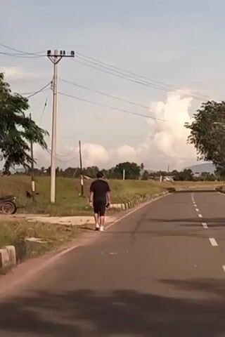 The video shows a person walking down a paved road in Buengpageu, Indonesia. The person, wearing a black t-shirt and black shorts, walks away from the camera on the right side of the road. They are walking alongside a grassy area with a concrete barrier and a utility pole with many wires. To the left of the road, there is a motorcycle parked on the grass. In the background, there are trees, fields, and some distant hills or mountains under a partly cloudy sky. The sun appears to be high in the sky, suggesting it is daytime, likely late morning or early afternoon. The lighting indicates a bright day, though the clouds suggest the possibility of scattered showers. The overall scene is rural and peaceful, with the person seemingly engaged in a leisurely walk. There is no visible text in the video.