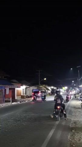 A night-time street scene with several motorcycles and cars. Two people are on a motorcycle in the foreground. In the background, other motorcycles and cars are driving on the road. There are buildings on the side of the road, and streetlights illuminate the scene. The overall impression is one of a busy street at night.